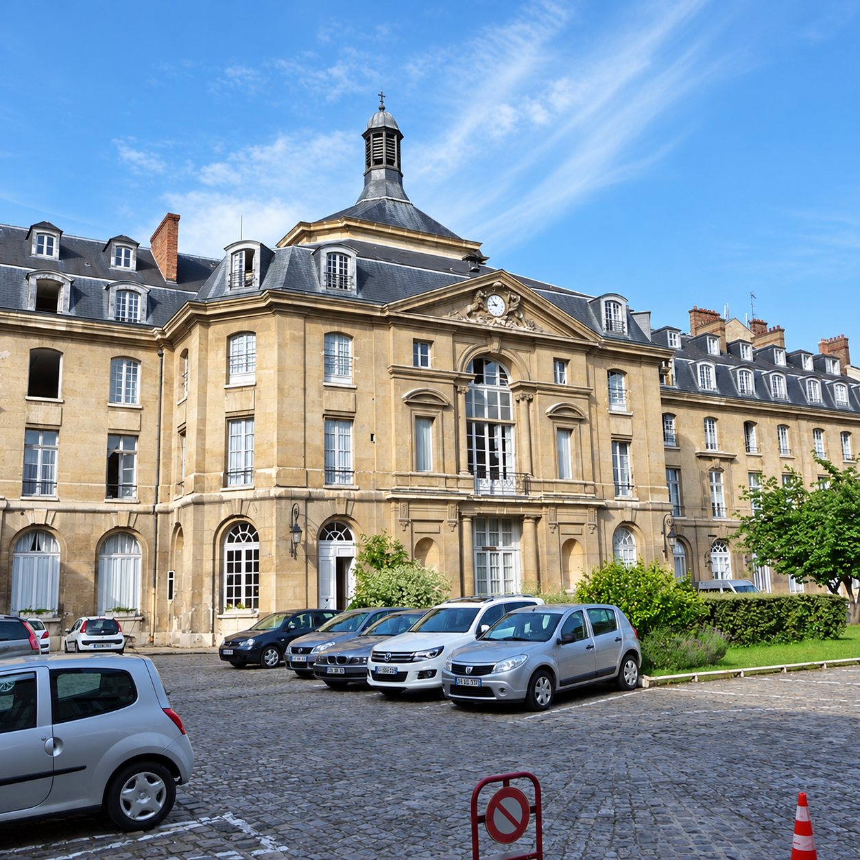 Façade d'un grand bâtiment ancien en pierre à toit d'ardoise et lanterneau, devant une cour pavée où plusieurs voitures sont stationnées.