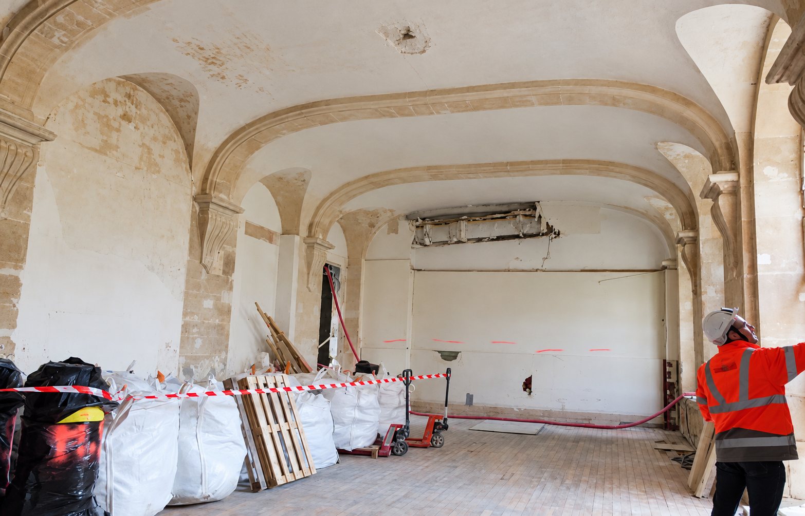 Un ouvrier en veste orange et casque blanc se tient dans une grande salle voûtée en pierre en rénovation, avec des big bags de gravats et des palettes alignés le long du mur.
