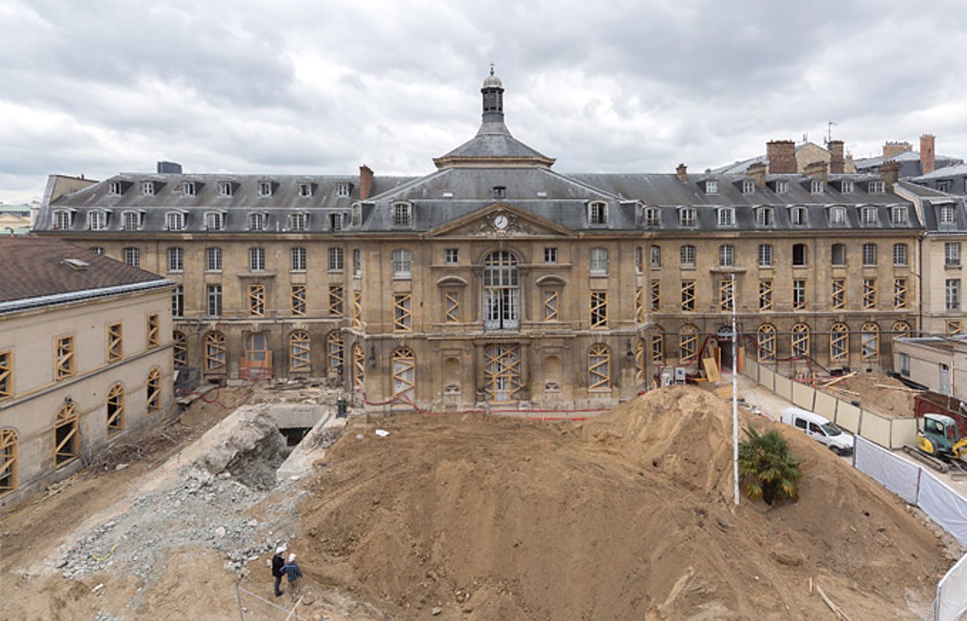 Un grand chantier de terrassement avec un monticule de terre et des gravats devant la façade d'un imposant bâtiment historique en pierre au toit d'ardoise, où deux ouvriers sont visibles.