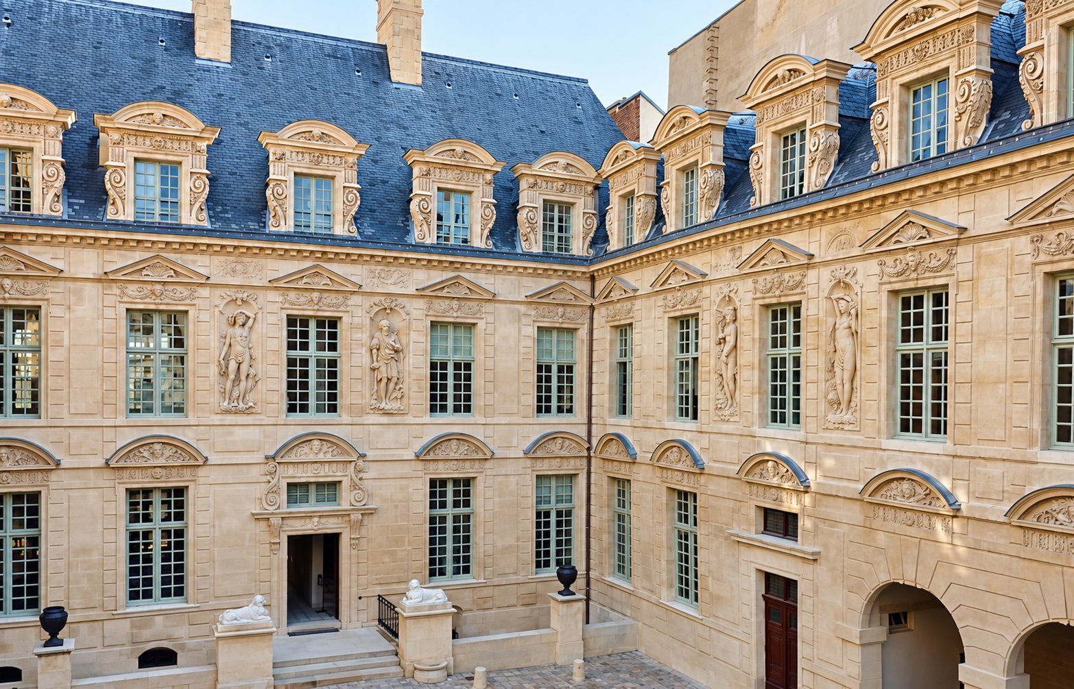 Façades en pierre de taille d'un hôtel particulier classique entourant une cour pavée, avec statues en bas-relief, hautes fenêtres et lucarnes ornées sur un toit d'ardoise.