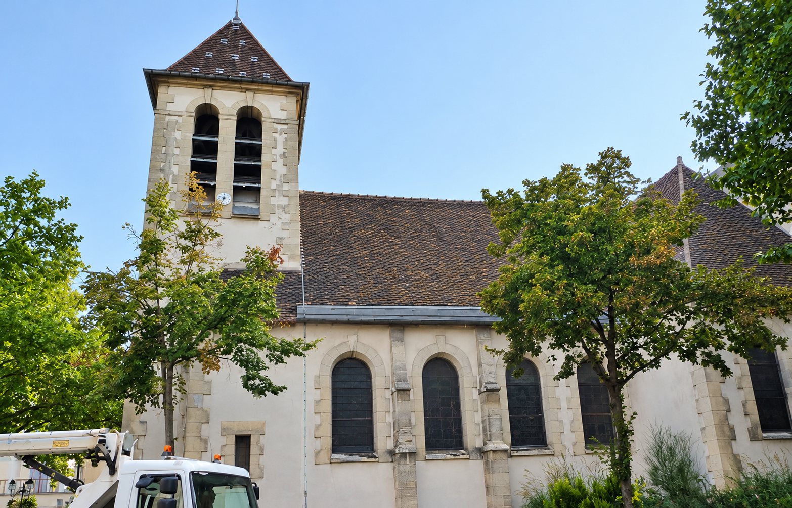 Une église en pierre claire avec son clocher et sa toiture en ardoise, entourée d'arbres, avec un camion équipé d'une nacelle visible en bas à gauche.