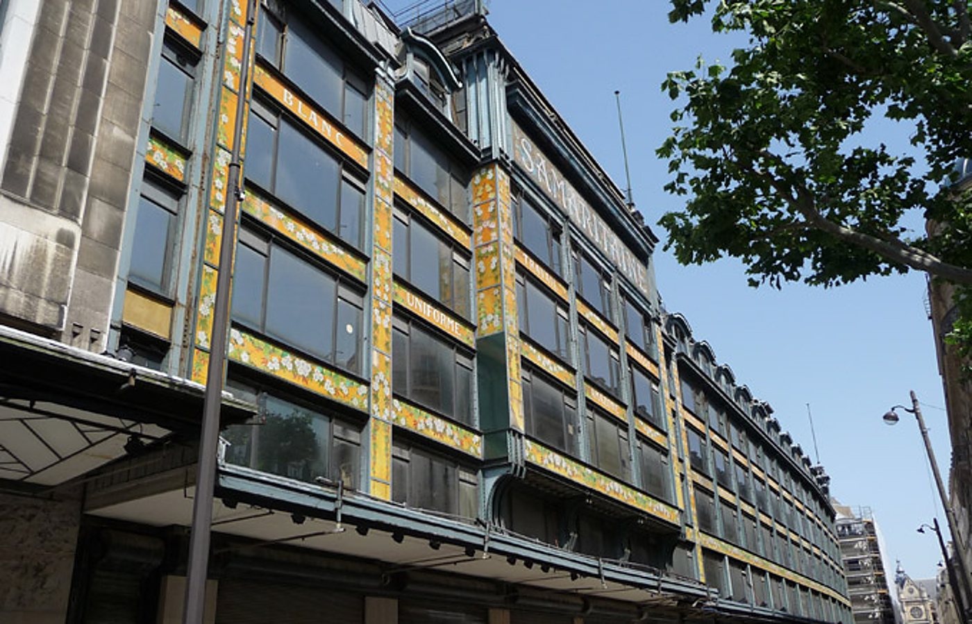 Façade d'un grand magasin de style Art nouveau, ornée de bandeaux dorés à motifs de fleurs, sous un ciel bleu et un arbre au premier plan.