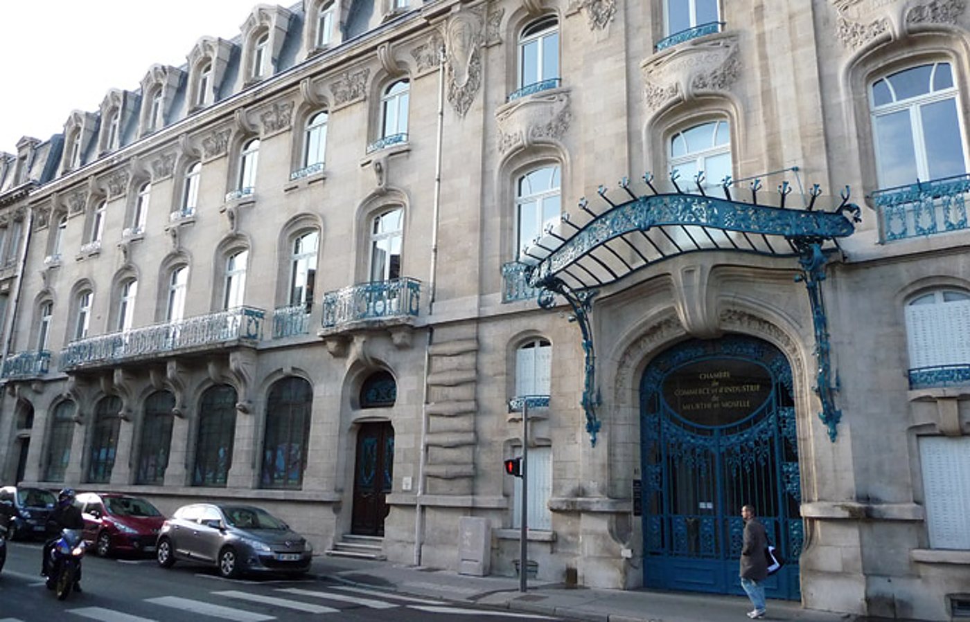 Façade d'un immeuble en pierre de style Art nouveau avec une grande porte et une marquise en fer forgé bleu, devant laquelle passent une voiture, une moto et un piéton.