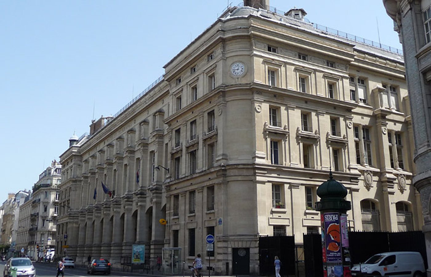Façade d'un grand immeuble en pierre avec une horloge, vu depuis l'angle d'une rue animée sous un ciel bleu, avec une colonne Morris au premier plan.