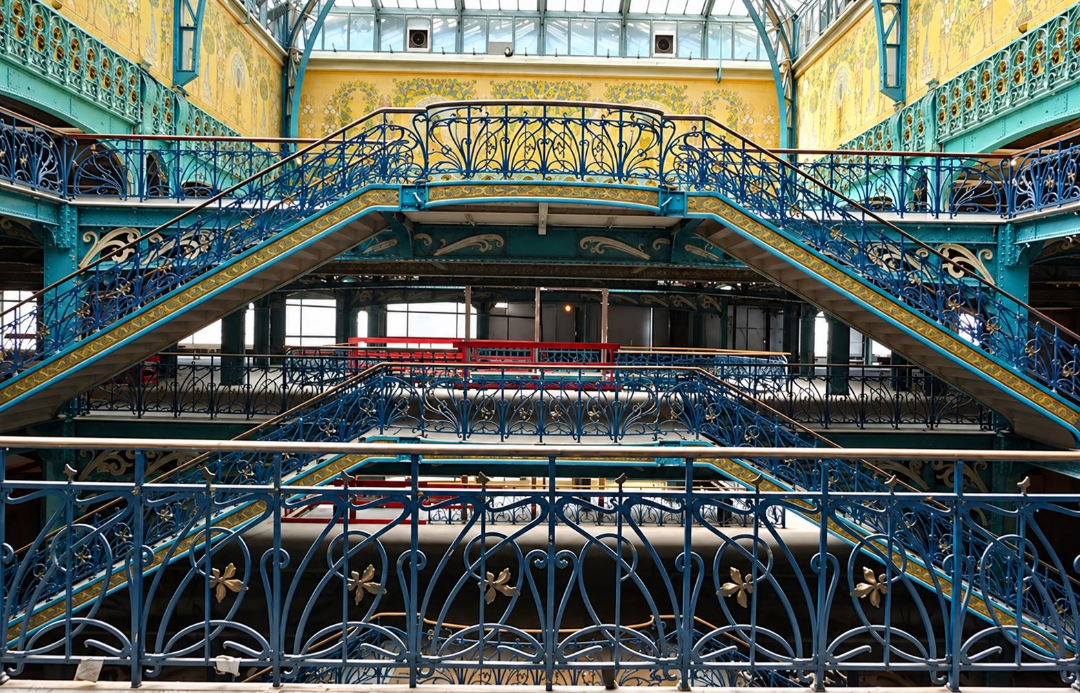 Intérieur d'un grand bâtiment avec escaliers symétriques à rampes en fer forgé bleu ornées de motifs Art nouveau, sous une verrière et des murs décorés.