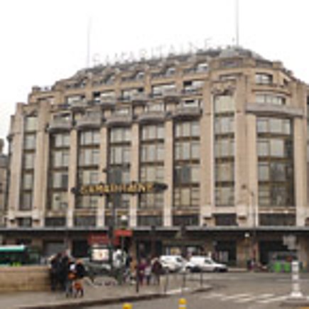Un grand immeuble de style haussmannien avec une façade en pierre et un toit mansardé, vu depuis la rue où passent des piétons et des voitures.