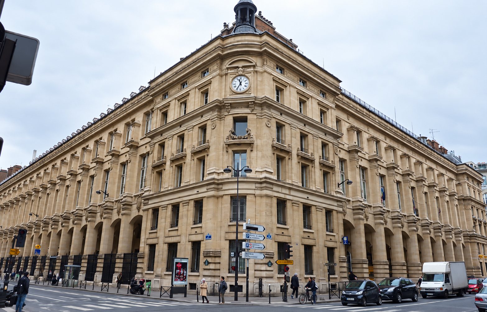 Un imposant bâtiment en pierre de style haussmannien avec une horloge sur la façade d'angle, entouré de rues avec voitures et piétons sous un ciel nuageux.