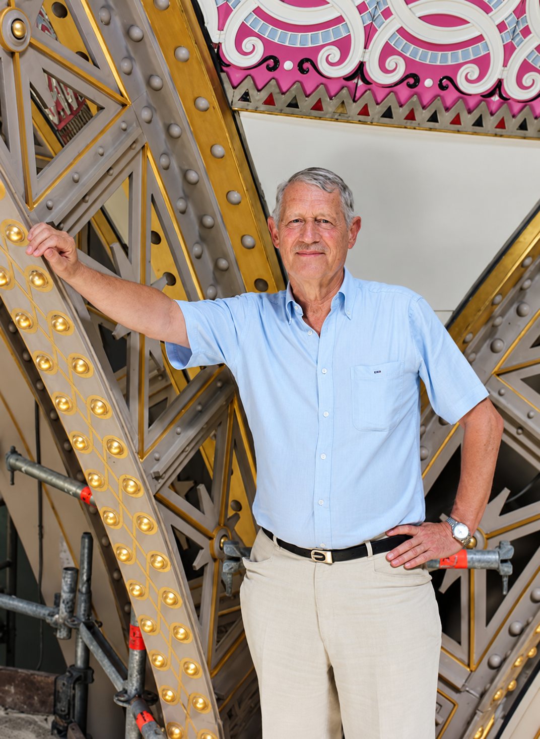 Un homme âgé aux cheveux gris, en chemise bleue claire et pantalon beige, pose la main sur une grande structure métallique rivetée sous un plafond décoré de motifs roses.
