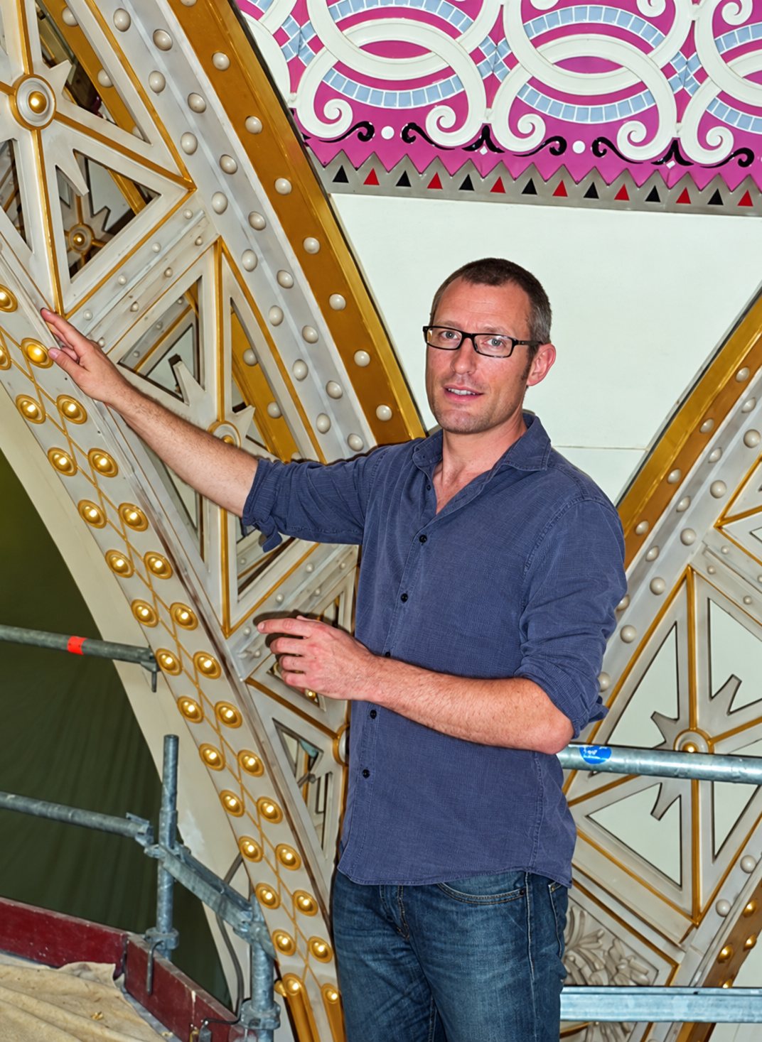 Un homme à lunettes en chemise bleue touche un décor ancien peint et doré, sur un échafaudage, pendant des travaux de restauration.