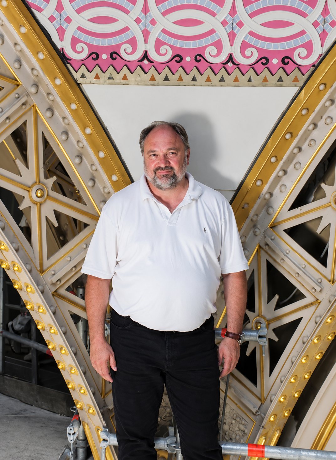 Un homme barbu en polo blanc et pantalon noir pose debout entre deux grandes arches métalliques dorées et ajourées, sous un décor peint rose et blanc.