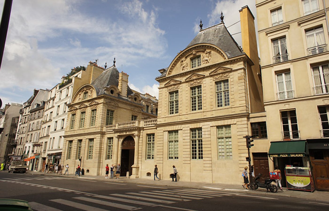 Façade en pierre d'un hôtel particulier du XVIIe siècle donnant sur une rue parisienne avec passants, passage piéton et scooter garé devant une pizzeria.