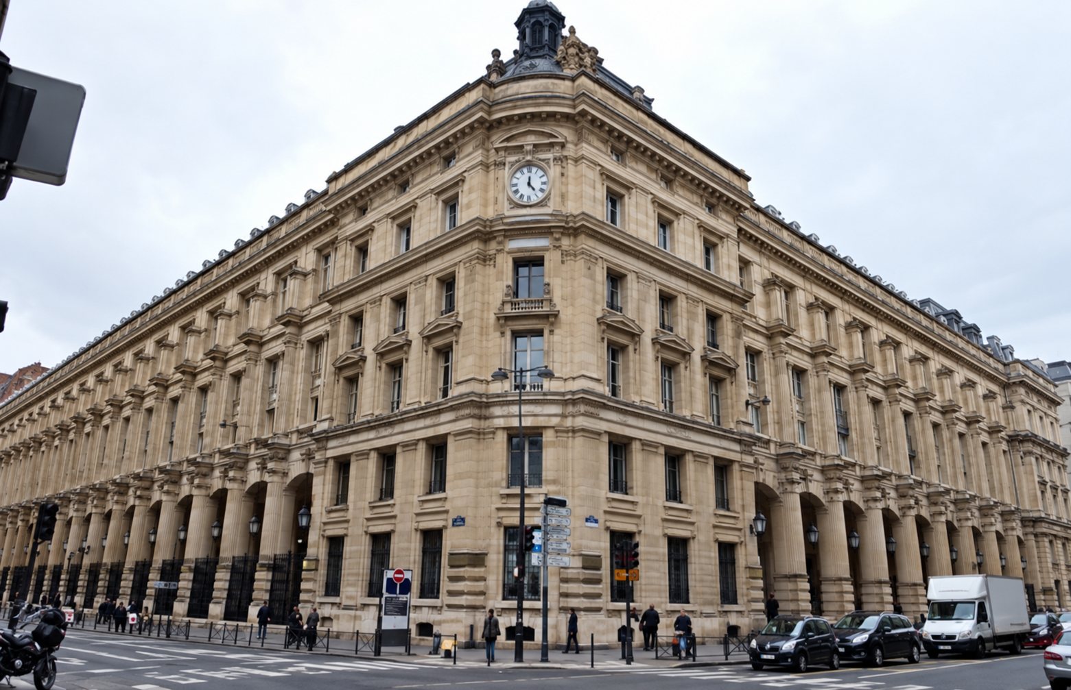Un grand bâtiment de pierre de style haussmannien, surmonté d'une horloge, se dresse à l'angle de deux rues animées sous un ciel gris.