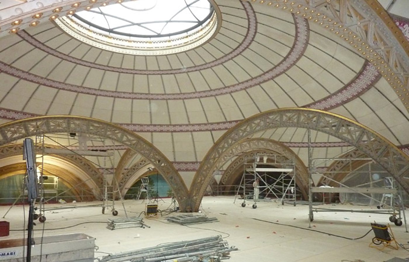 Intérieur d'une vaste salle à coupole décorée de motifs, en cours de rénovation, avec des échafaudages, des arches métalliques dorées et du matériel posé au sol.