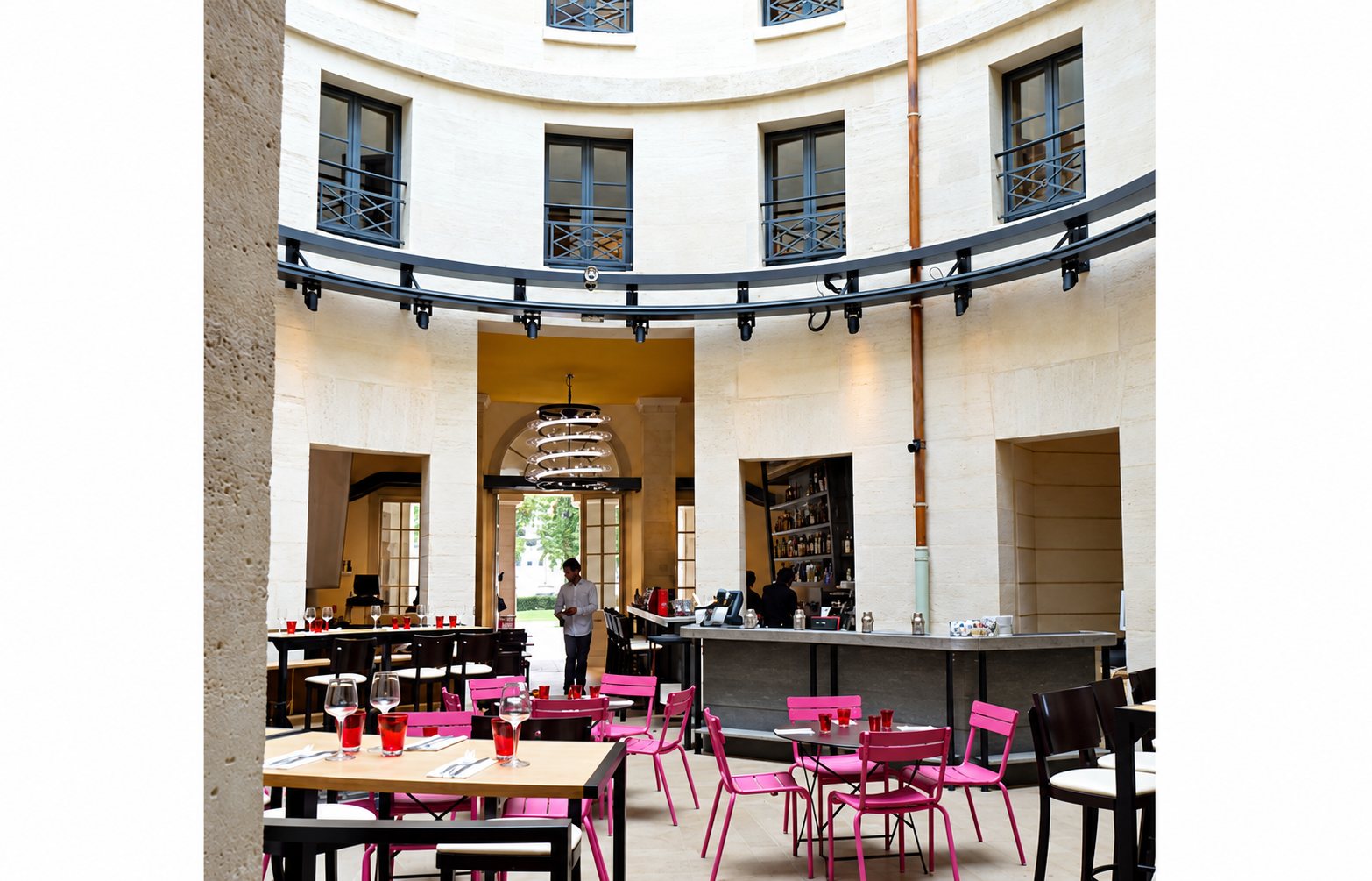 Salle de restaurant installée dans une cour circulaire en pierre claire, avec des tables en bois, des chaises roses et un comptoir de bar au fond.