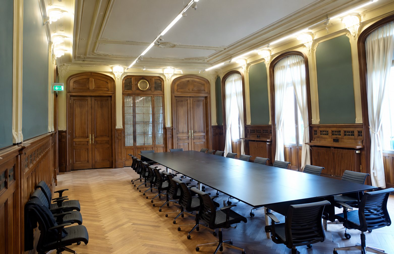 Une grande salle de réunion au parquet en chevrons, aux murs vert-gris et boiseries, meublée d'une longue table noire entourée de fauteuils de bureau.