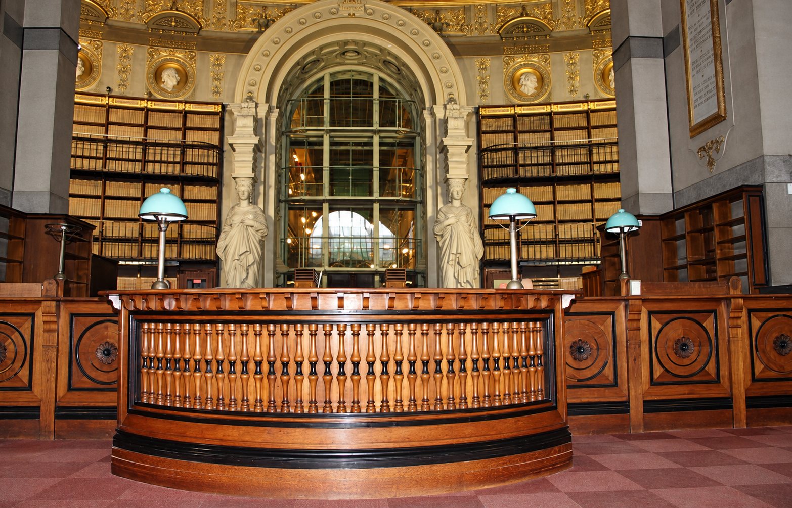 Une grande salle de lecture ancienne avec une banque circulaire en bois à balustres, des lampes de bureau vertes, des rayonnages de livres et une grande verrière en arrière-plan.