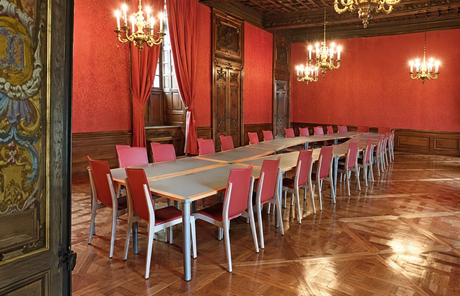 Une grande salle de réception aux murs rouges et lustres dorés, meublée d'une longue table blanche entourée de chaises rouges sur un parquet ancien.