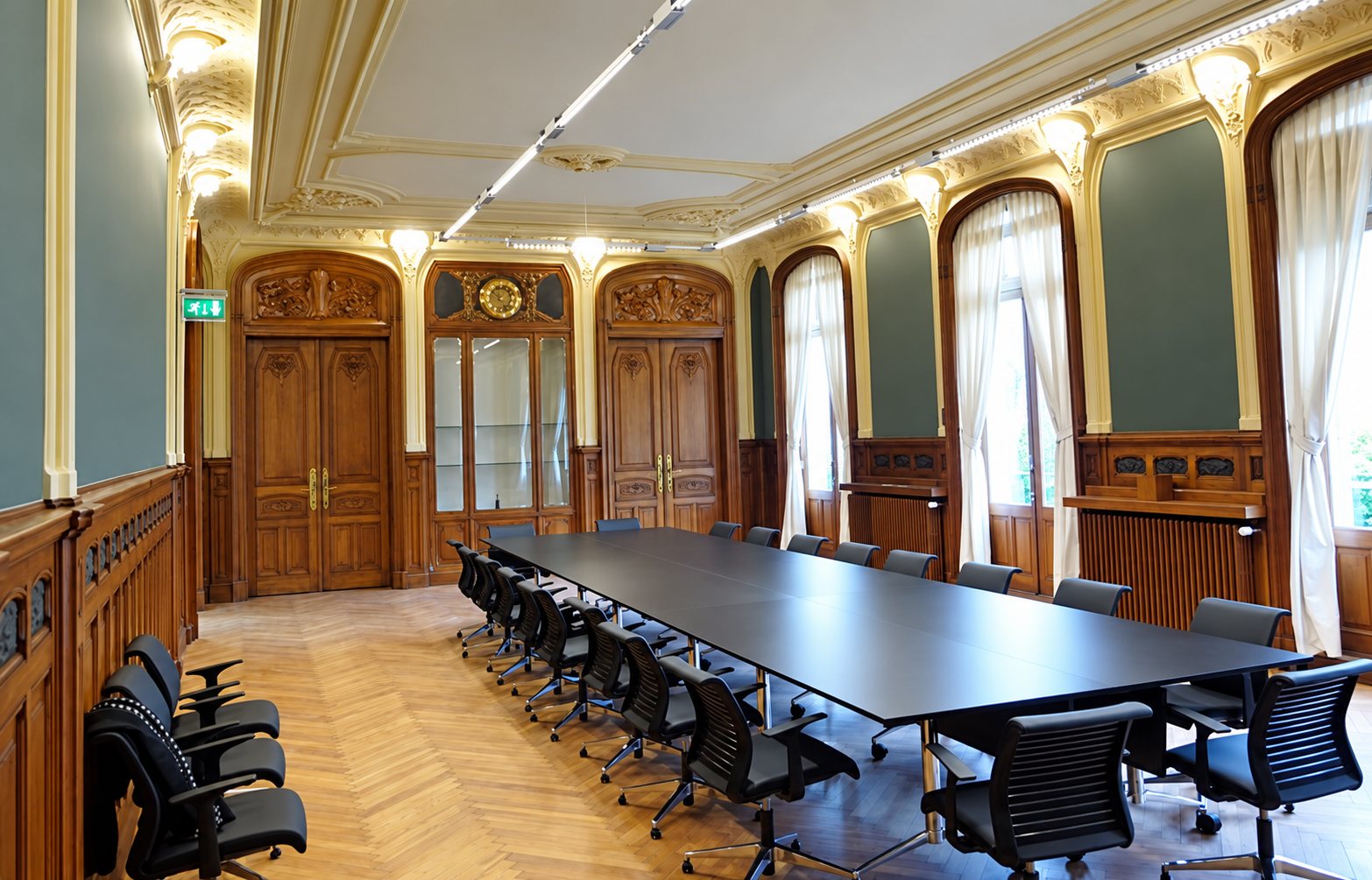 Une grande salle de réunion historique avec parquet en chevrons, boiseries sombres, hautes fenêtres drapées et une longue table noire entourée de fauteuils.