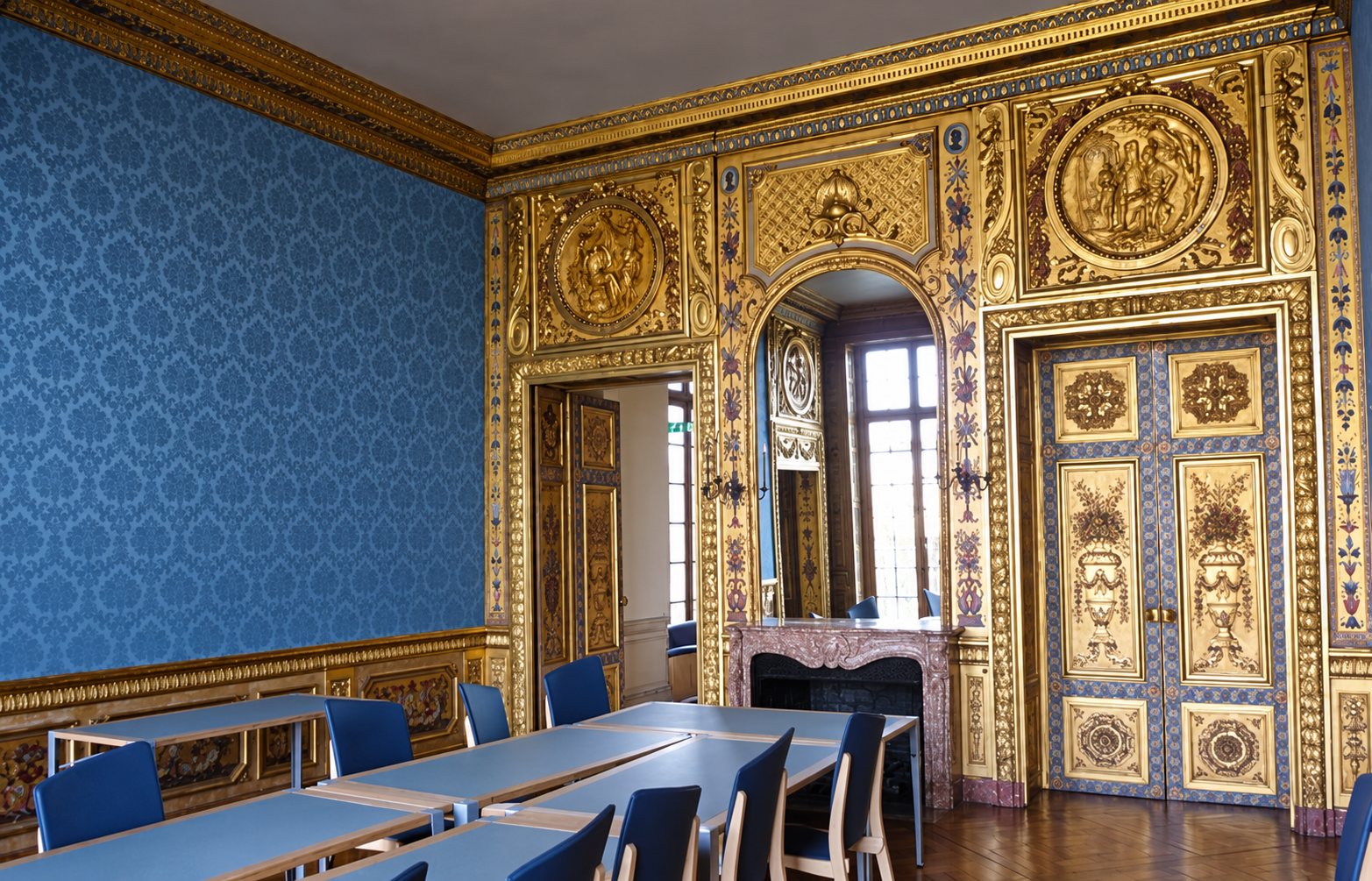Une salle ancienne aux murs tendus de tissu bleu et aux riches boiseries dorées, meublée de tables grises et de chaises bleues devant une cheminée en marbre.