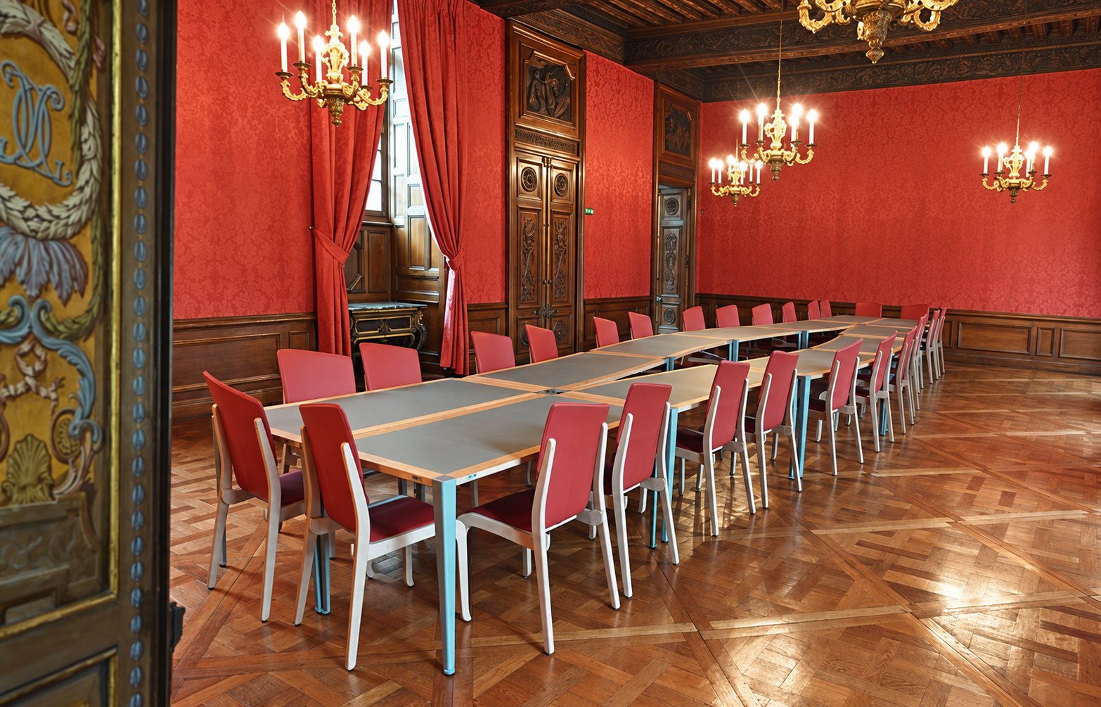 Une salle de réunion ancienne aux murs rouges damassés, au parquet ciselé et aux lustres, meublée d'une longue table modulable et de chaises rouges.