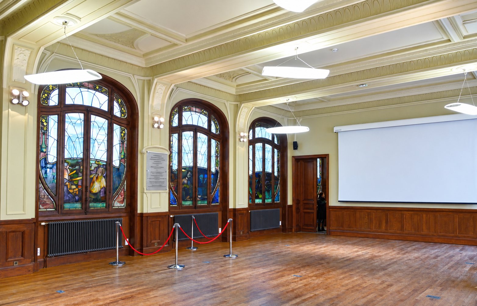 Une grande salle au parquet ancien avec des vitraux Art nouveau, des lampes suspendues, des cordons rouges et un écran de projection blanc.
