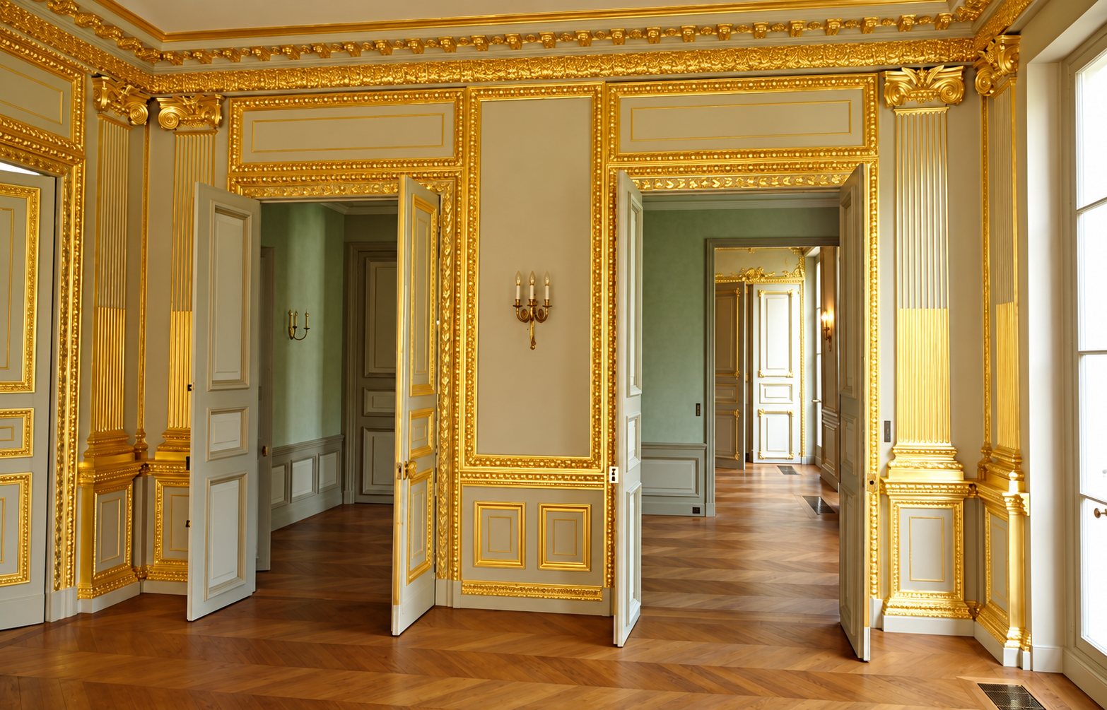 Pièce de style classique avec boiseries grises rehaussées de dorures, portes ouvertes sur d'autres salons et parquet en chevrons.