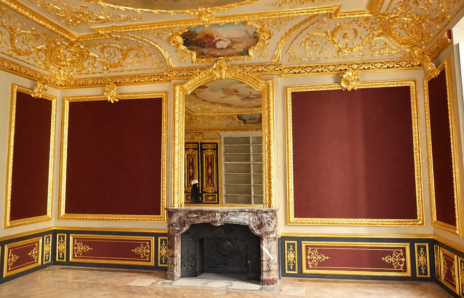 Salon de style classique aux boiseries dorées et panneaux bordeaux, avec une cheminée en marbre surmontée d'un grand miroir et d'un plafond peint.