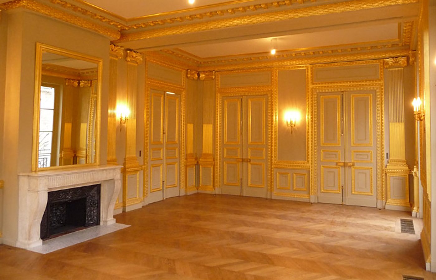 Grand salon ancien aux boiseries grises rehaussées de dorures, avec cheminée en marbre blanc, grand miroir doré et parquet en chevrons.