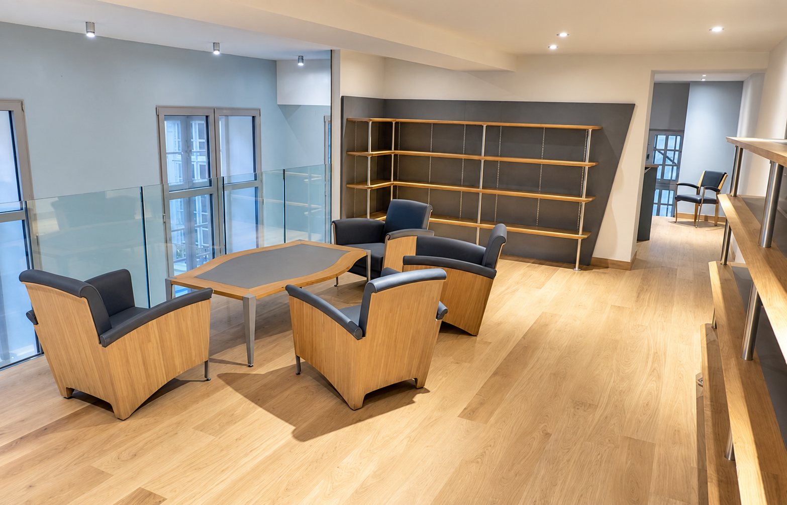Un salon lumineux au parquet clair, avec quatre fauteuils en bois et cuir gris autour d'une table basse, des étagères en bois et une balustrade en verre.