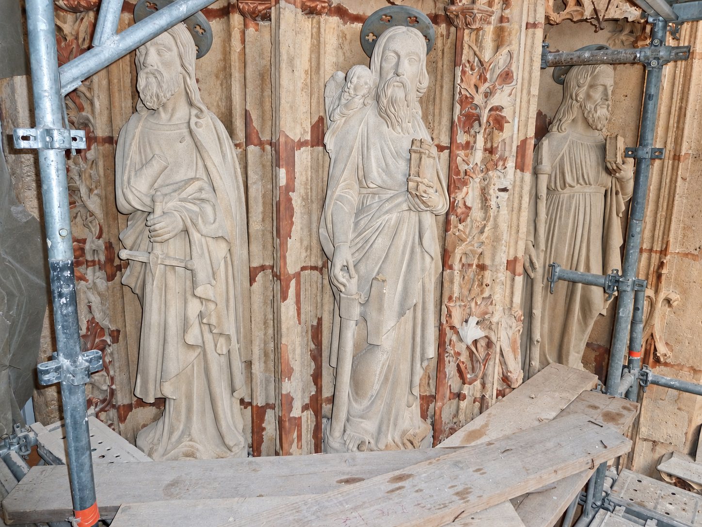 Trois statues de saints en pierre avec auréoles, entourées d'un échafaudage métallique et de planches, pendant un chantier de restauration.