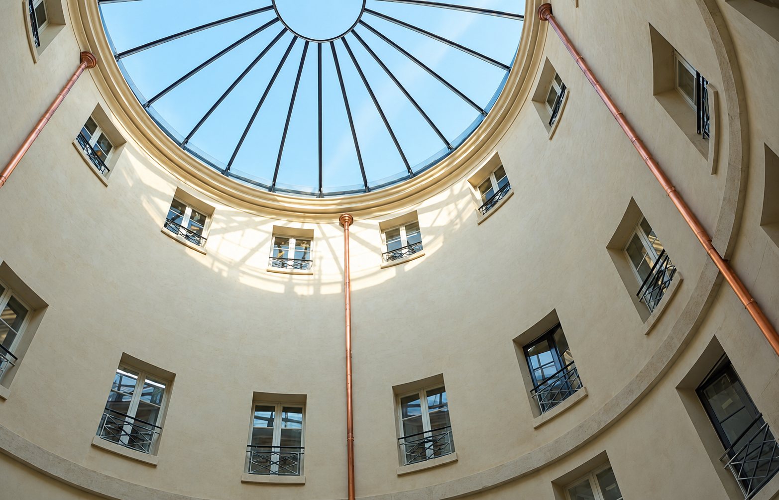 Vue en contre-plongée d'une cour circulaire aux murs clairs, percée de fenêtres à garde-corps et surmontée d'une grande verrière, avec des descentes en cuivre.